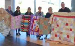 People holding colorful quilts in a bright room, standing in a row.