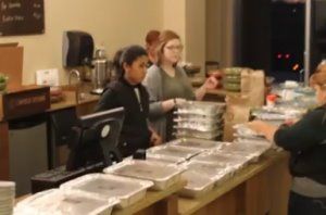 Volunteers serving food behind a long counter with trays in a community kitchen or cafeteria