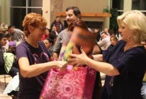 Two women hand a pink gift bag to a child in a busy indoor gathering.