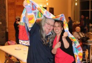 Two smiling people holding up a colorful quilt in a banquet hall