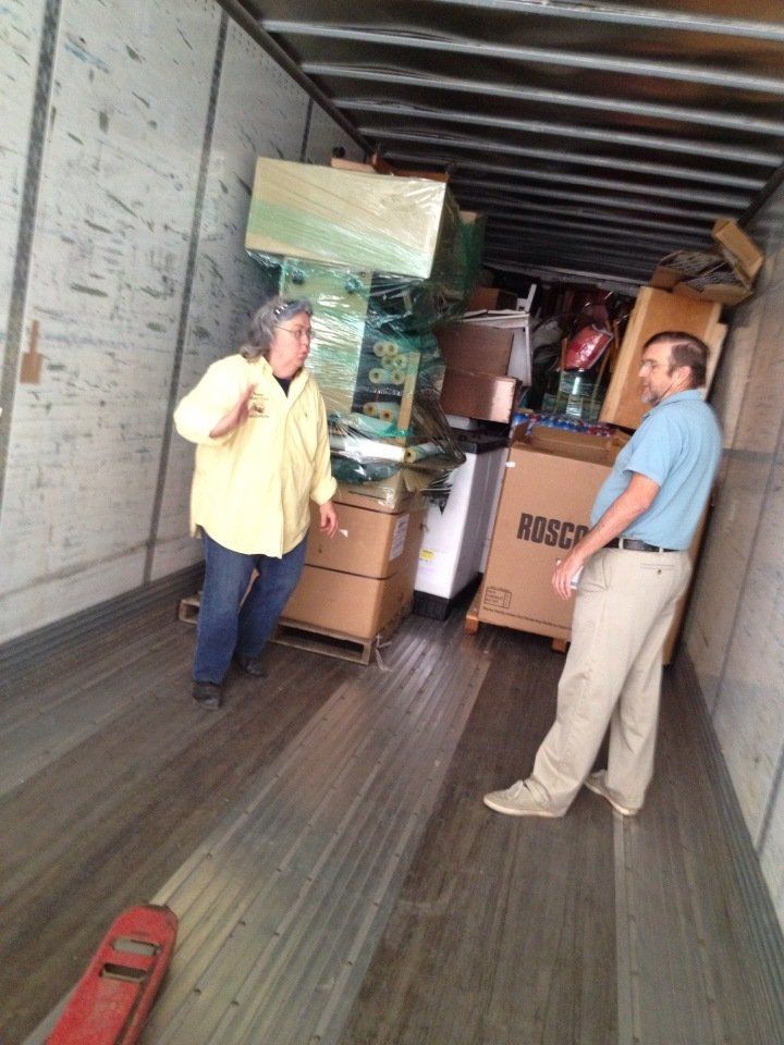Two men stand in a loaded moving truck surrounded by stacked boxes and furniture.