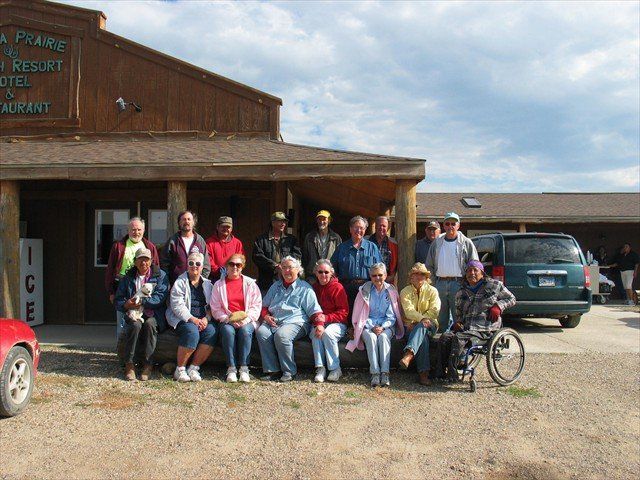 Group posing outside a rustic lodge with parked vehicles and bicycles on a gravel lot