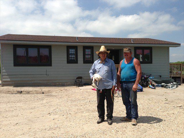 Two people standing in front of a single-story house on a gravel lot.