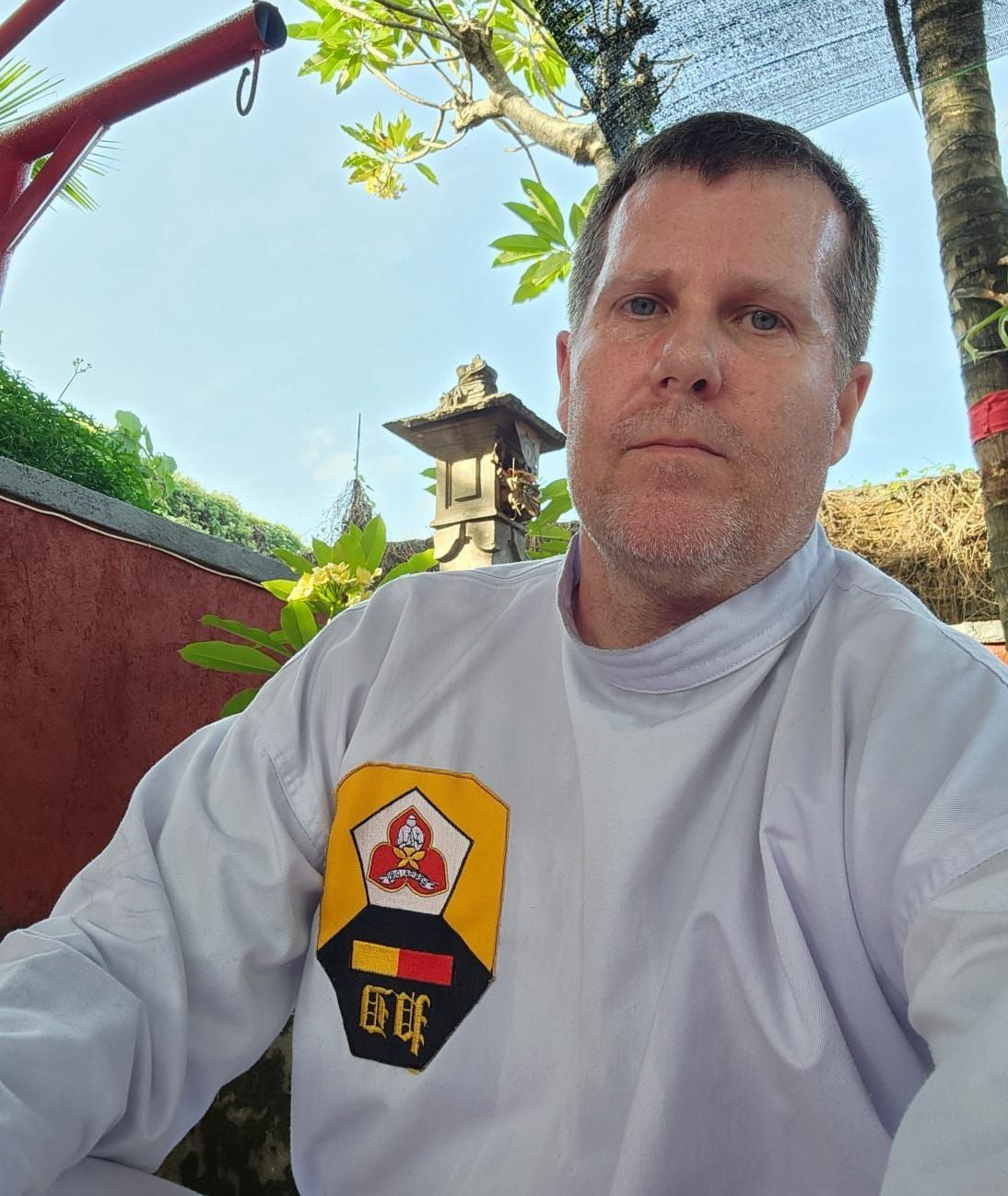 A man wearing a white shirt with a yellow patch on his chest is taking a selfie.