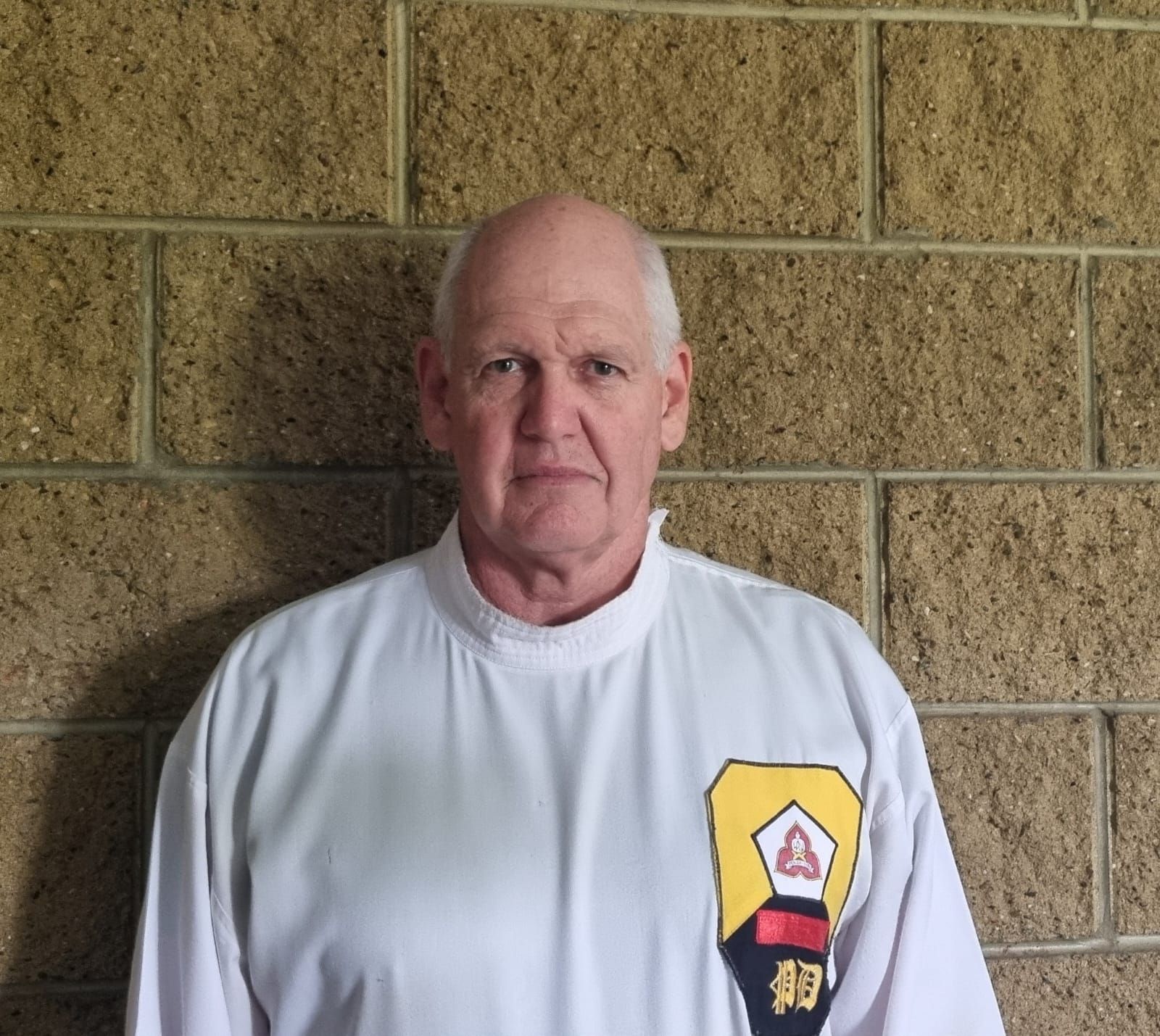 A man wearing a white shirt with a yellow patch on his chest is standing in front of a brick wall