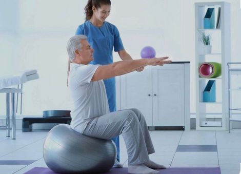 Un hombre mayor se ejercita sobre una pelota de estabilidad con un terapeuta en una luminosa sala de rehabilitación.