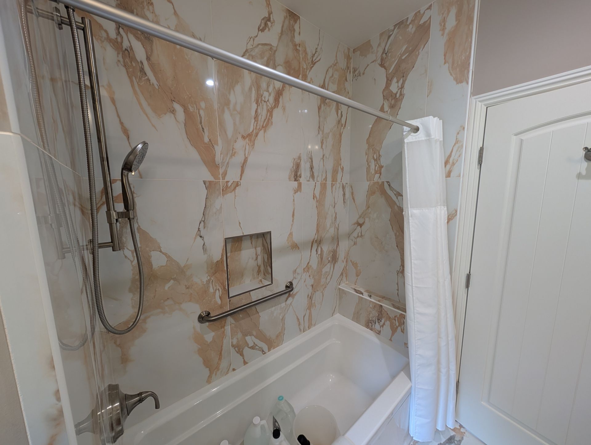Bathroom with marble-look walls, bathtub, handheld shower, and towel hanging on a grab bar.