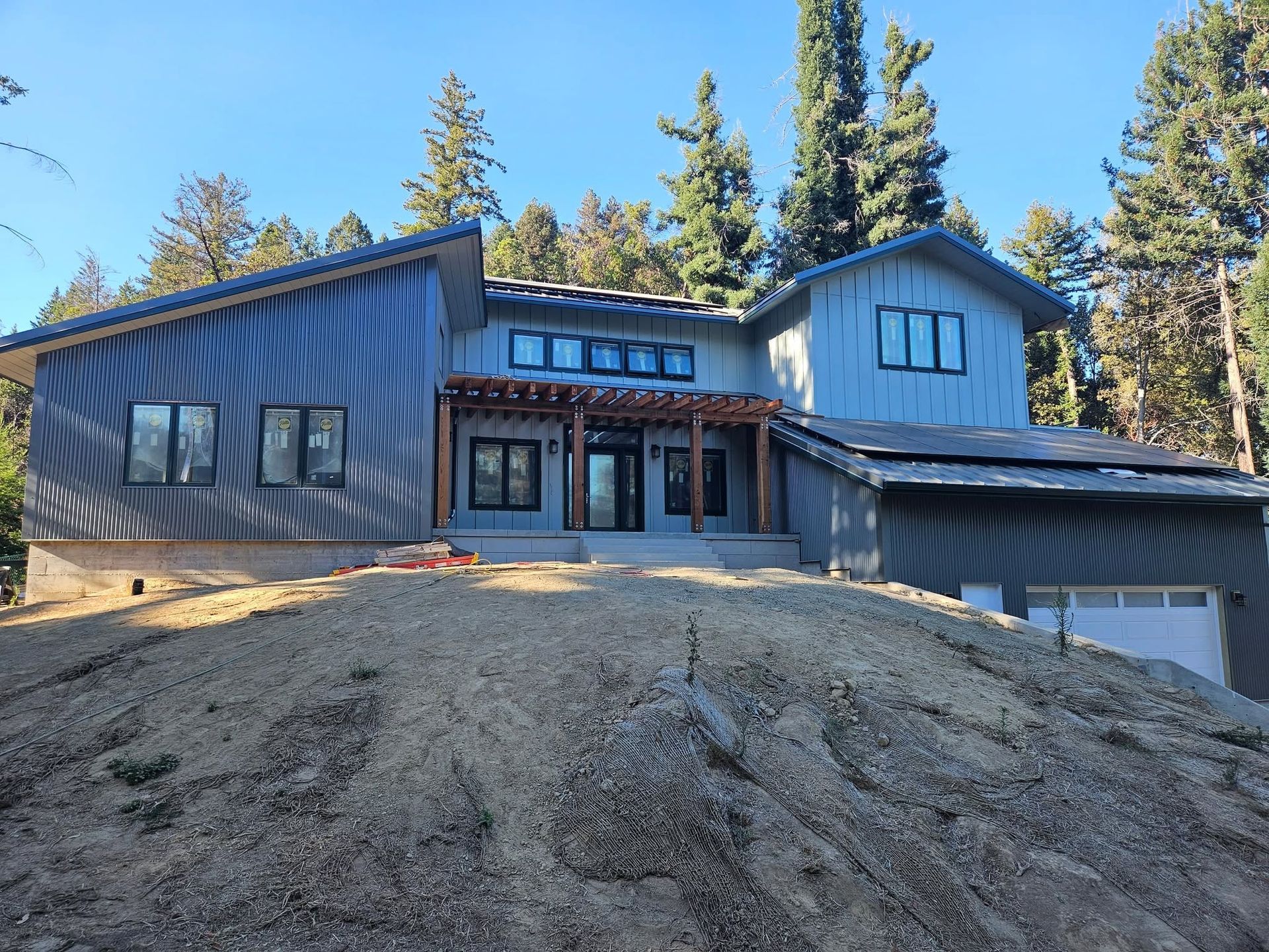 Modern blue-gray house on a dirt hillside with a large garage and tall trees under a clear sky