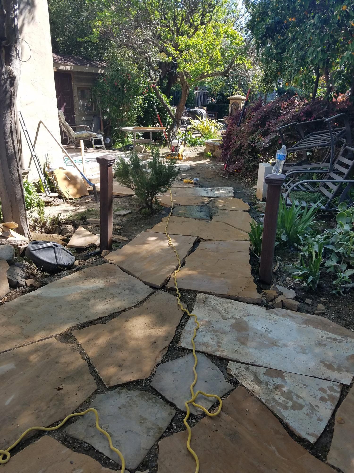 Broken stone garden path with cracks, leaning posts, and overgrown greenery in a backyard.
