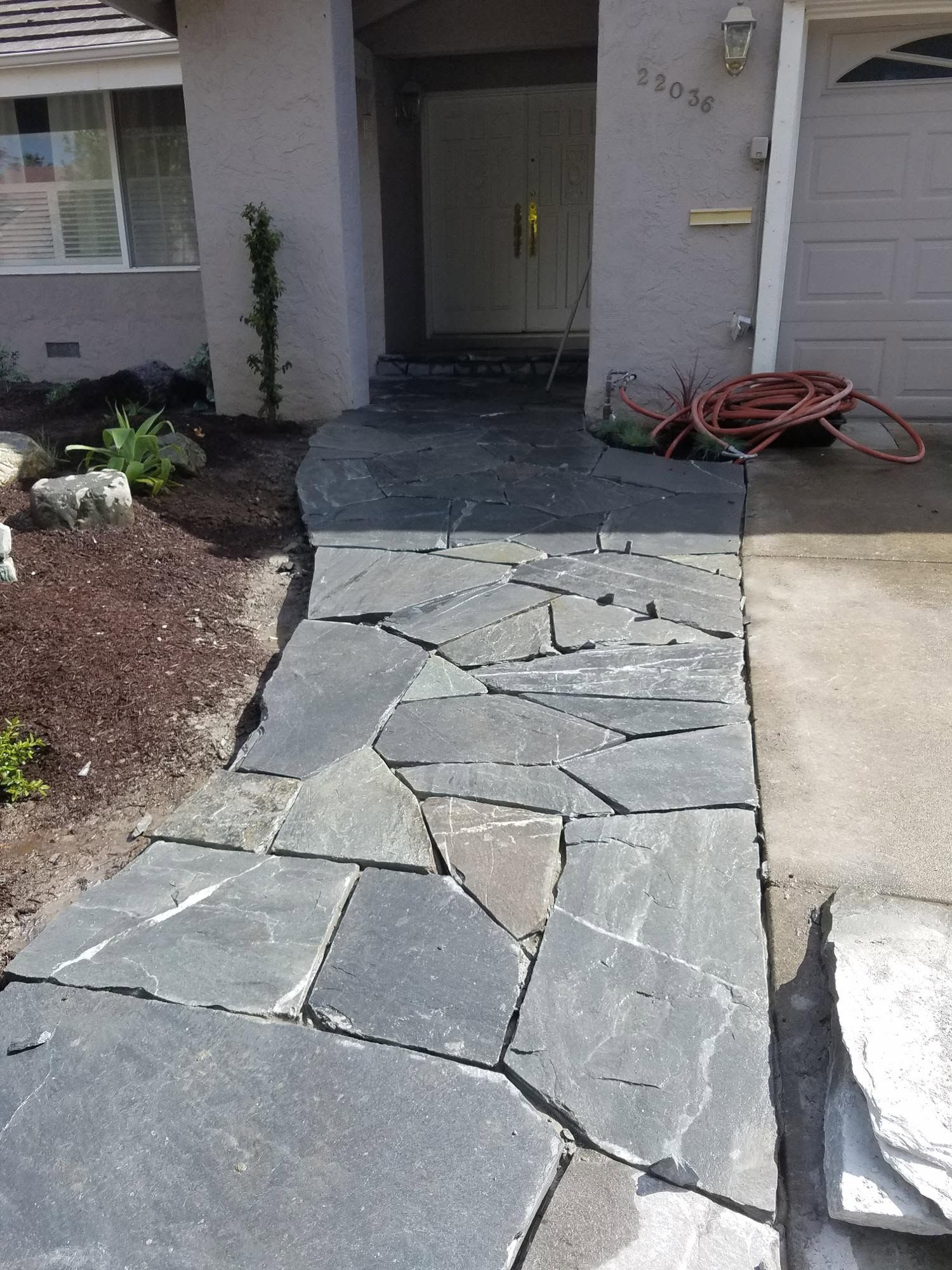 Damaged stone walkway leading to a front door, with broken pavers and a red hose on the right