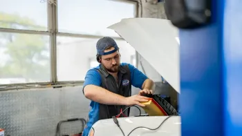 Mechanic working under a raised car hood in a garage, holding a tool near the engine