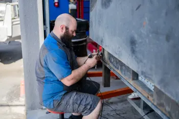 Mechanic kneeling beside a truck, working with tools under the chassis in a garage.