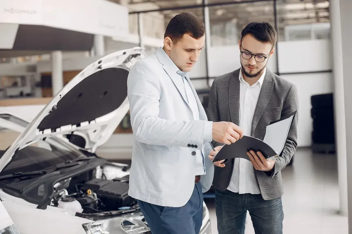 Two mechanics inspect a car engine and discuss a tablet in an auto garage