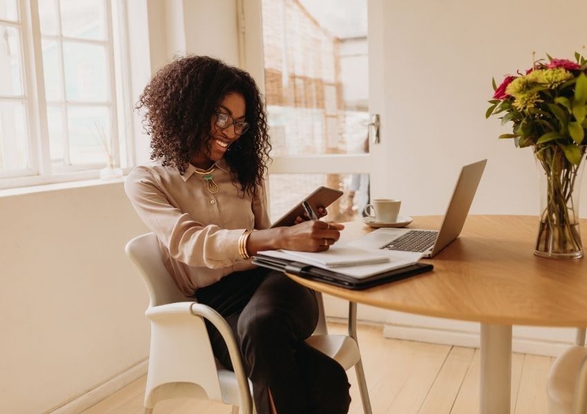 Woman working at a wooden table with a laptop and coffee, holding a phone in a bright room