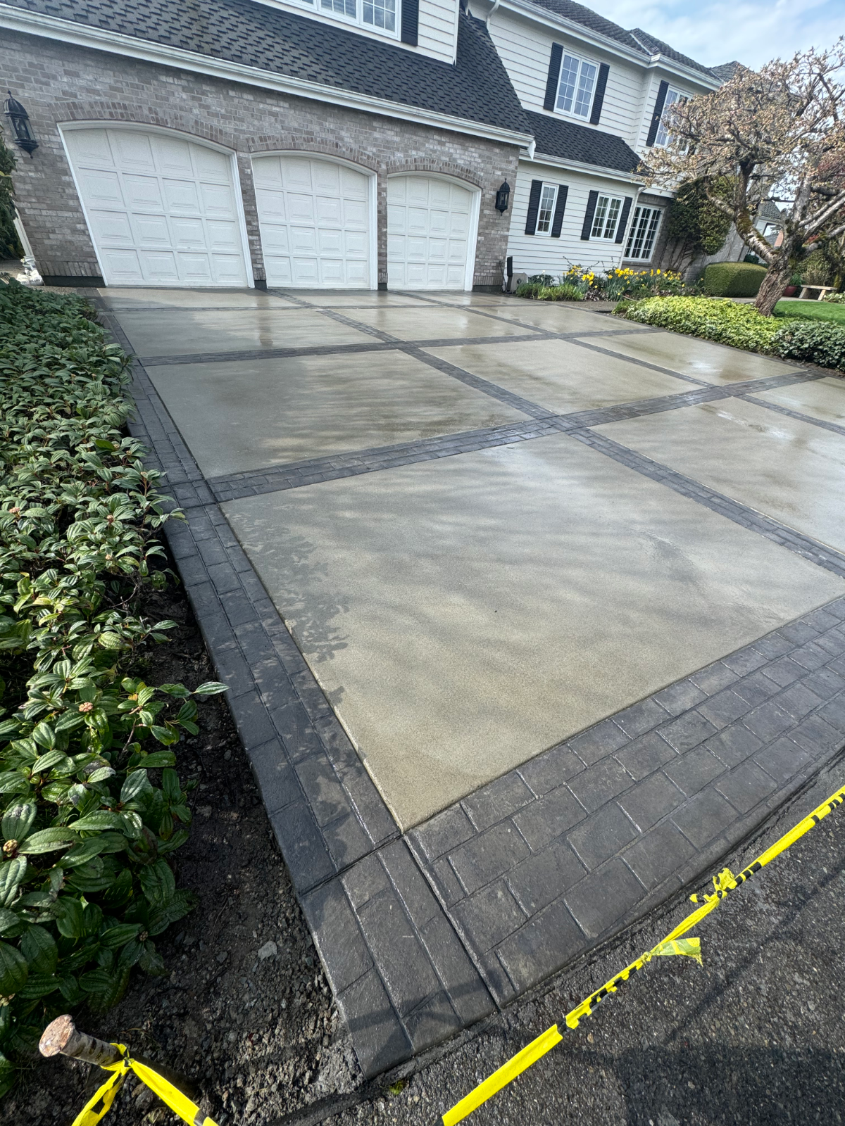 Wet concrete driveway with two garage doors, dark brick border, and shrubs along the side