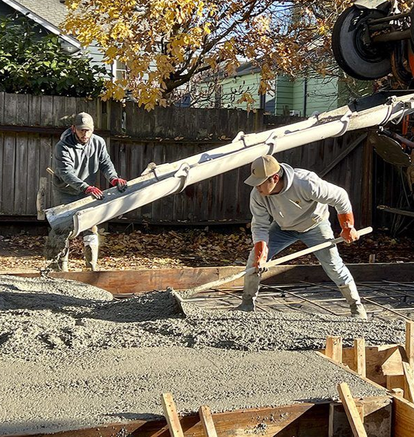 Two workers pouring and leveling concrete at a construction site using a long chute.