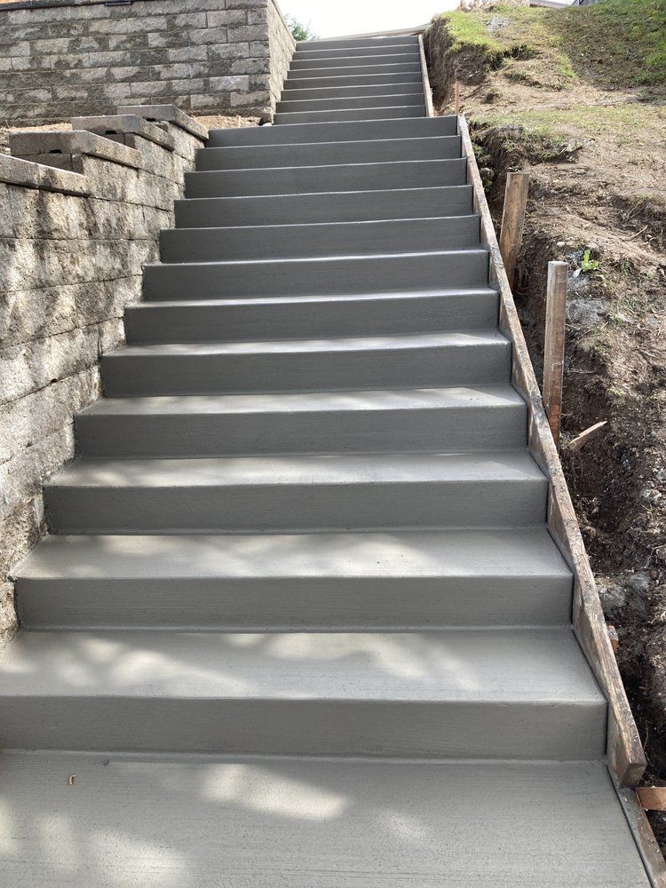 Concrete staircase outdoors beside a stone wall and dirt embankment, leading uphill.
