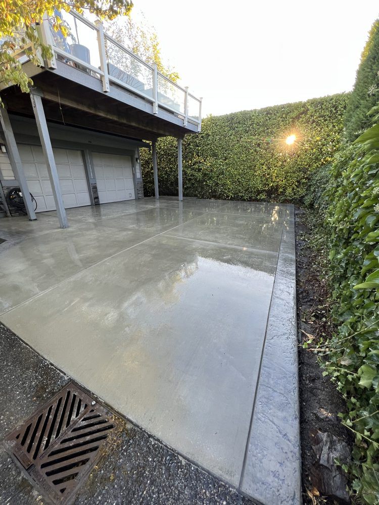 Wet concrete patio beside a raised deck, bordered by tall hedges and a drain grate in the foreground
