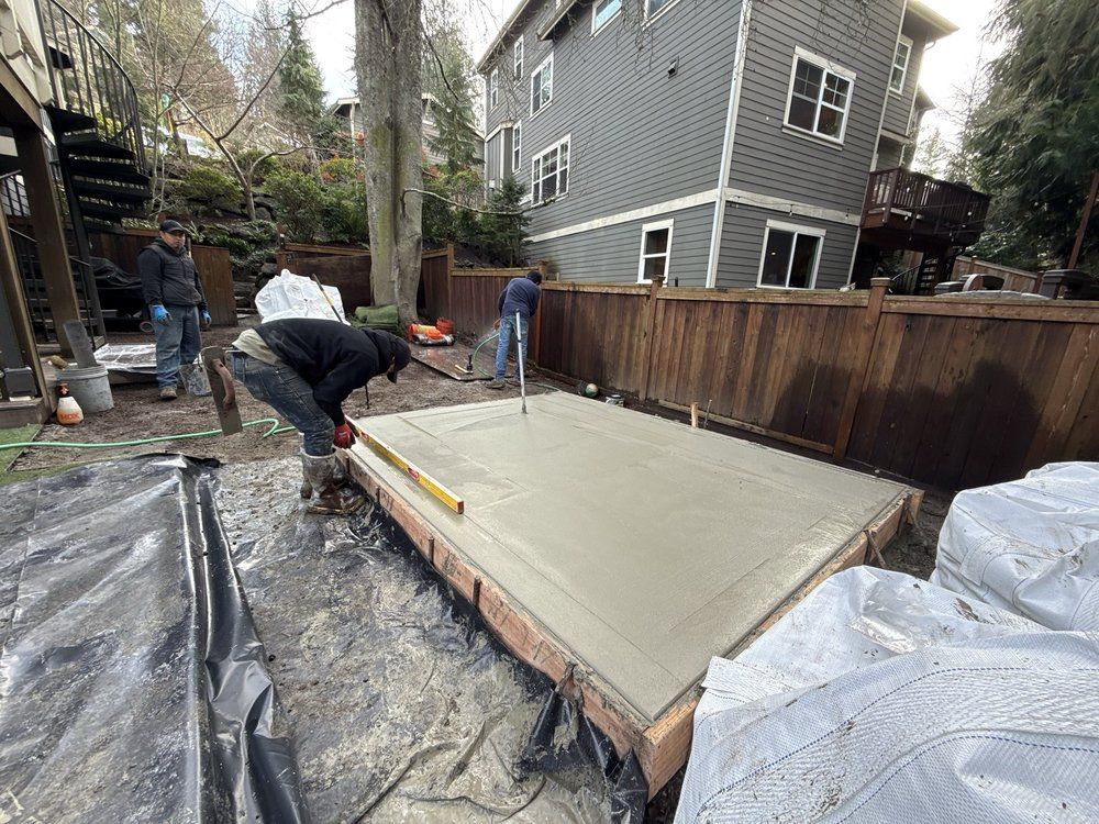 Workers pouring a concrete patio slab in a backyard beside a wooden fence and house.