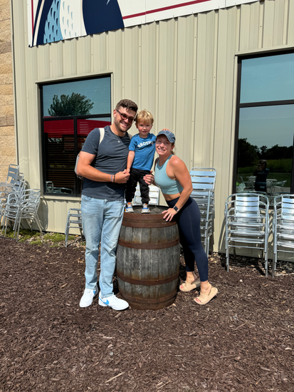 A man, woman, and toddler are standing next to a barrel in front of a building.