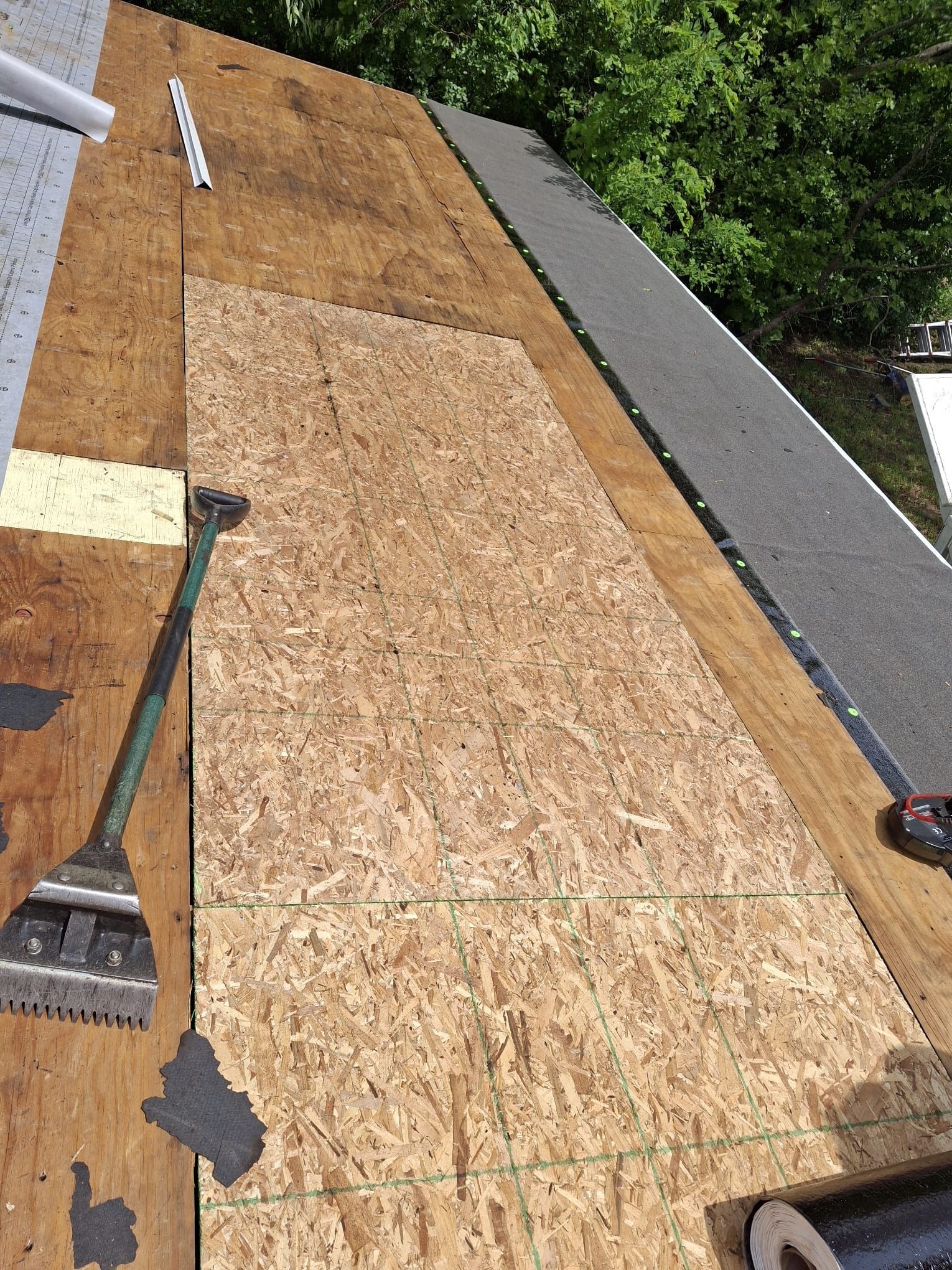 Partially shingled roof with exposed plywood decking and tools along the edges