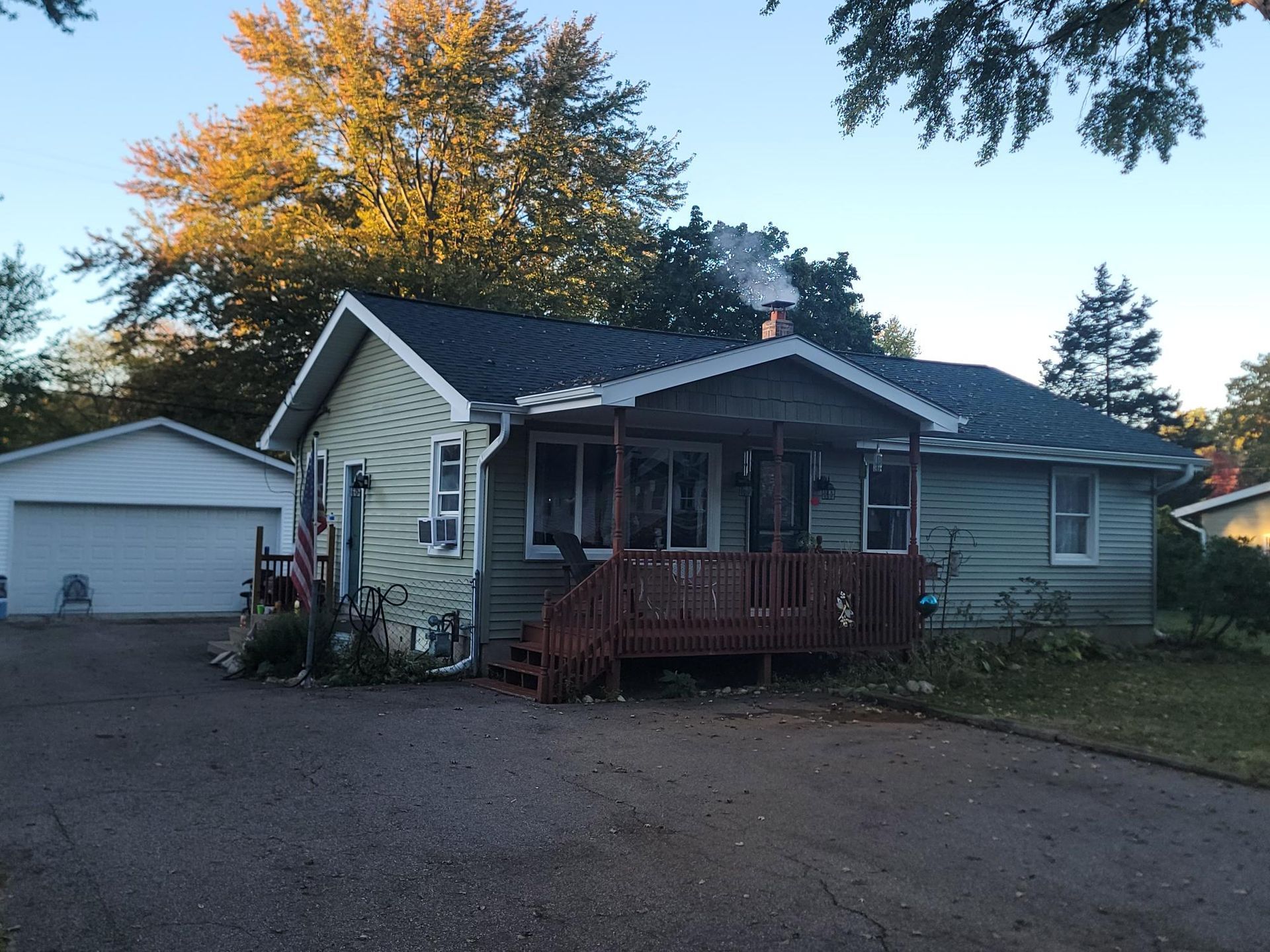 Small beige house with a red porch, detached garage, and autumn trees at dusk
