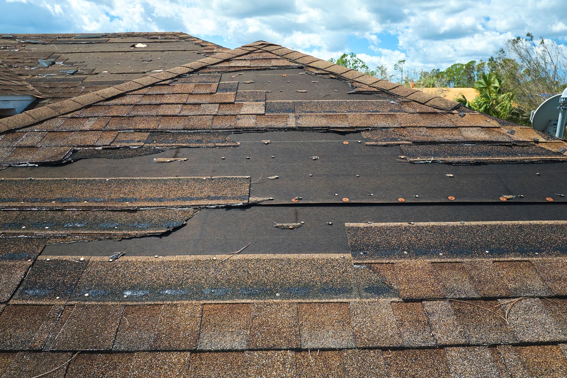 Damaged shingled roof with missing and curled tiles under a blue sky