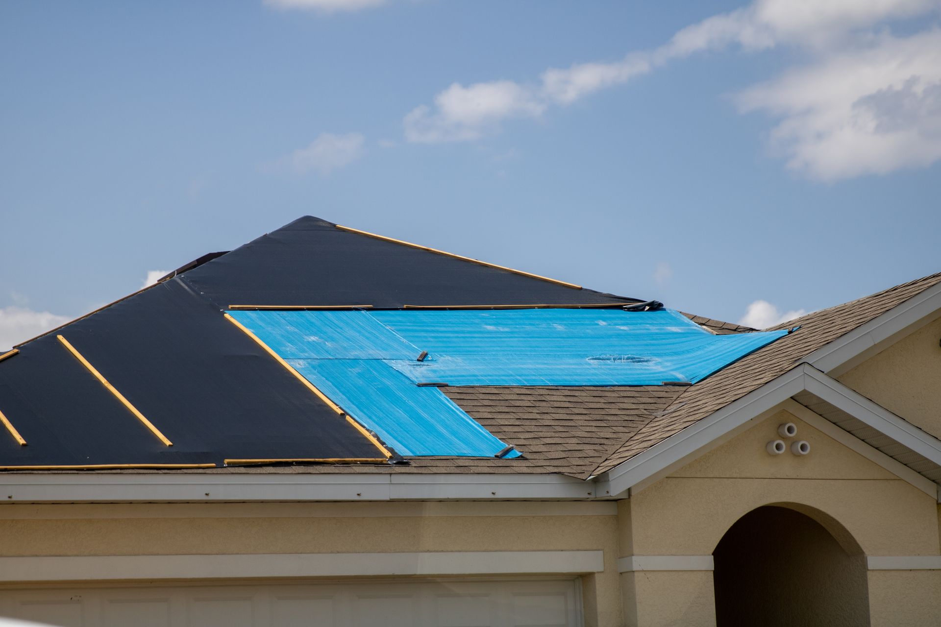 House roof under construction with black underlayment and blue protective covering under a partly cloudy sky