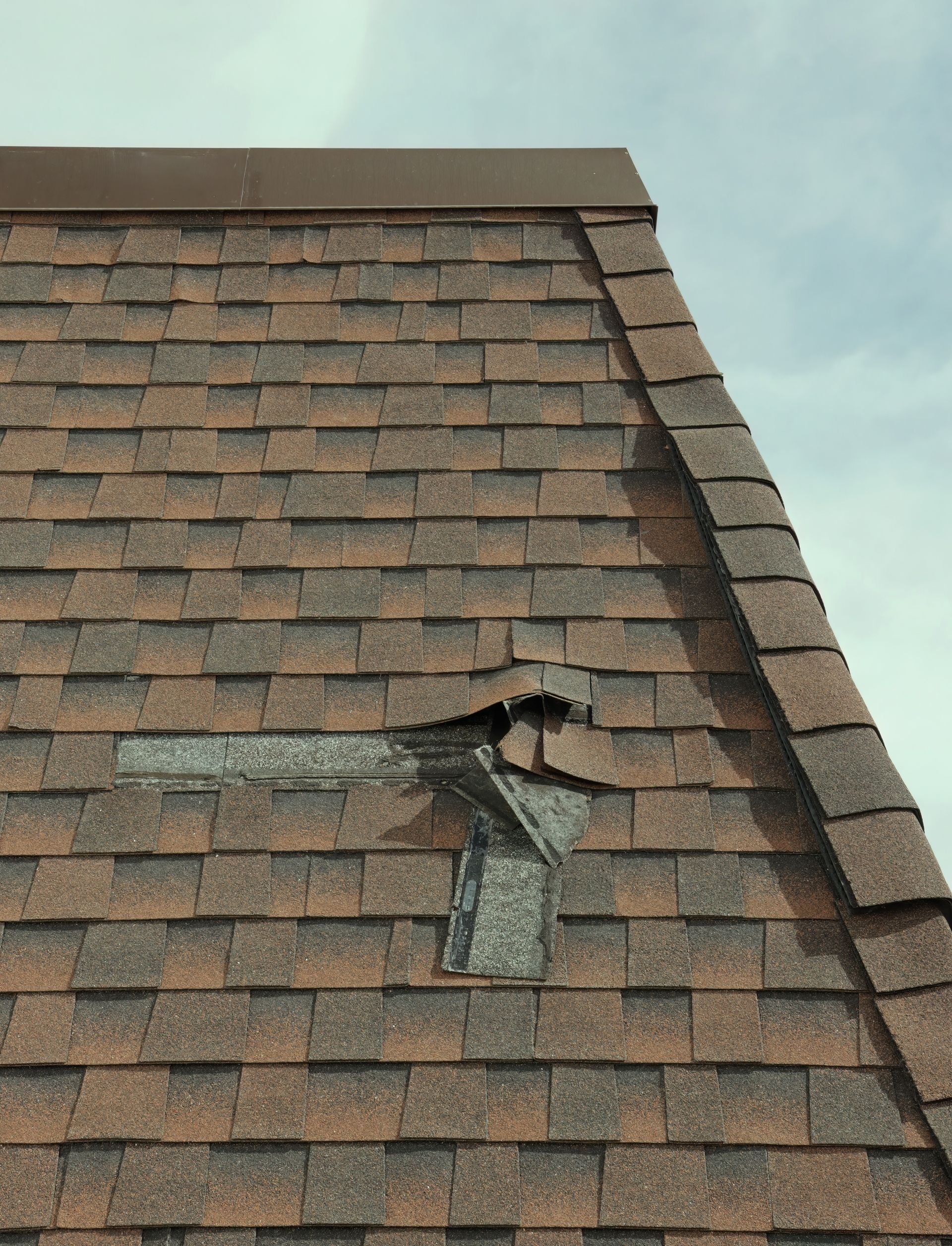 Damaged shingled roof with a torn vent and visible hole under cloudy sky