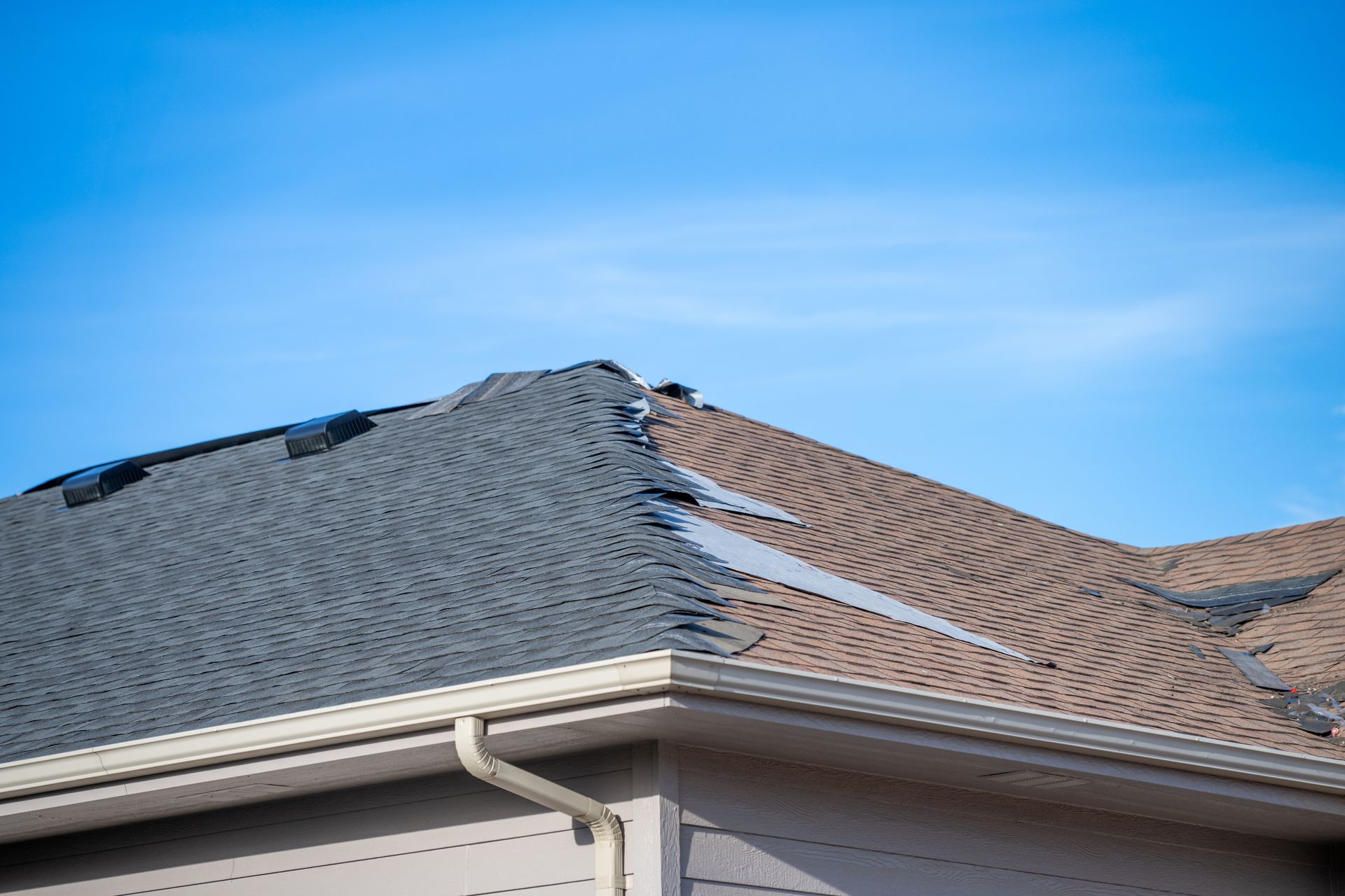 Damaged gray shingle roof with missing sections beneath a blue sky