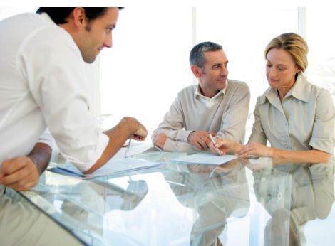 Tres personas están sentadas alrededor de una mesa de cristal revisando documentos, inmersas en una consulta profesional en una oficina luminosa.