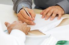 Primer plano de unas manos escribiendo en un documento con un bolígrafo plateado, sentadas en un escritorio.