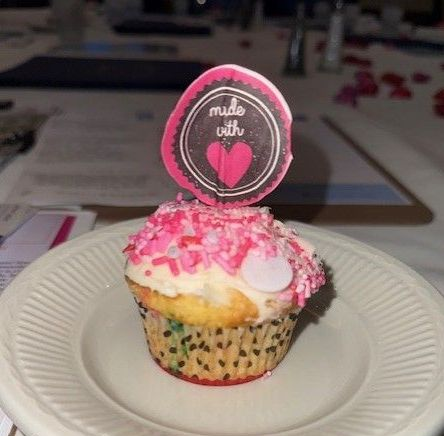 A vanilla cupcake with white frosting and pink sprinkles sits on a white plate, topped with a 
