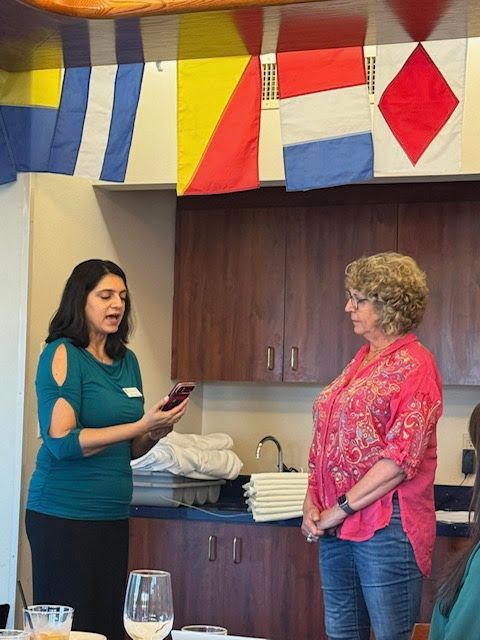 A person in a teal top reads from a phone to a person in a pink top in a room with nautical flags hanging above.