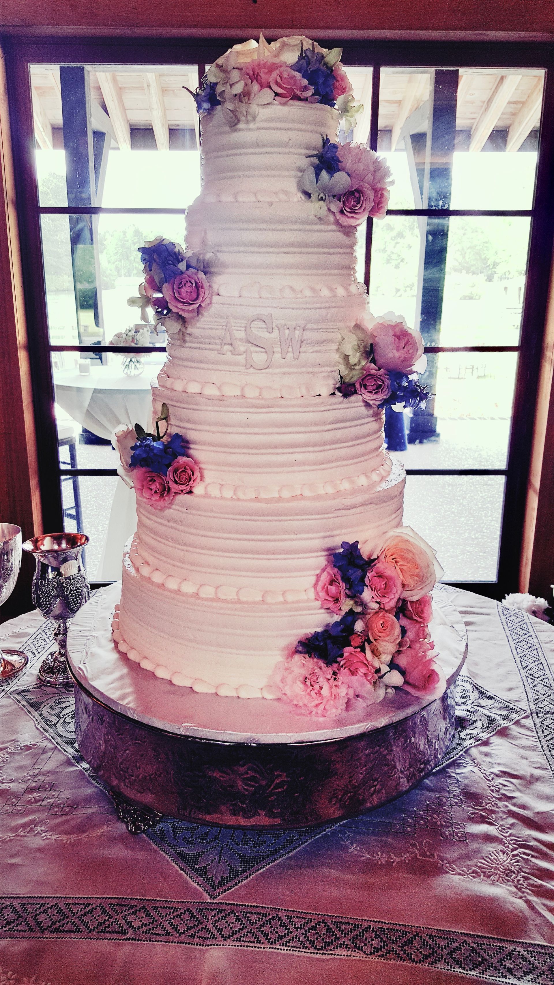 Tall white tiered wedding cake with pink and purple flowers on a wooden table by a window