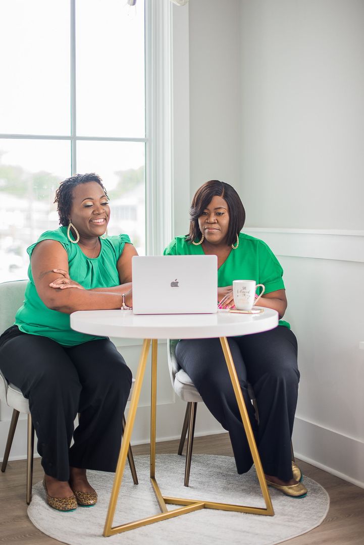 Two women in green tops sit at a round table using a laptop in a bright room.
