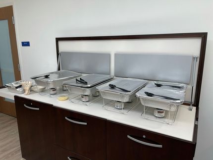 Self-serve buffet with covered stainless-steel food trays on a dark wood counter.
