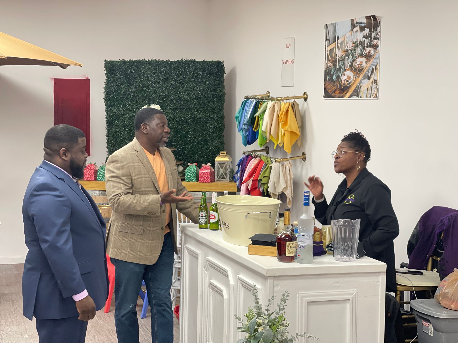 Three people stand at a bar counter in a colorful indoor event space, talking and holding drinks.