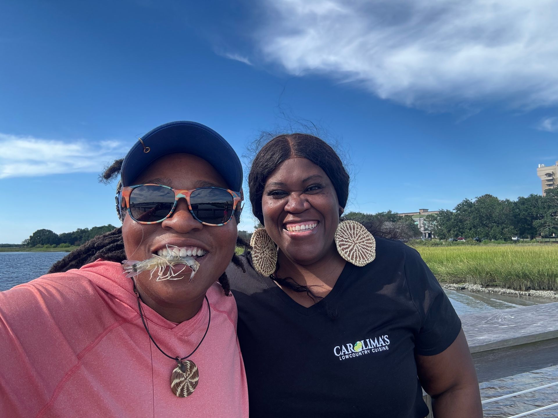 Two smiling friends posing outdoors on a sunny waterfront with blue sky and water behind them