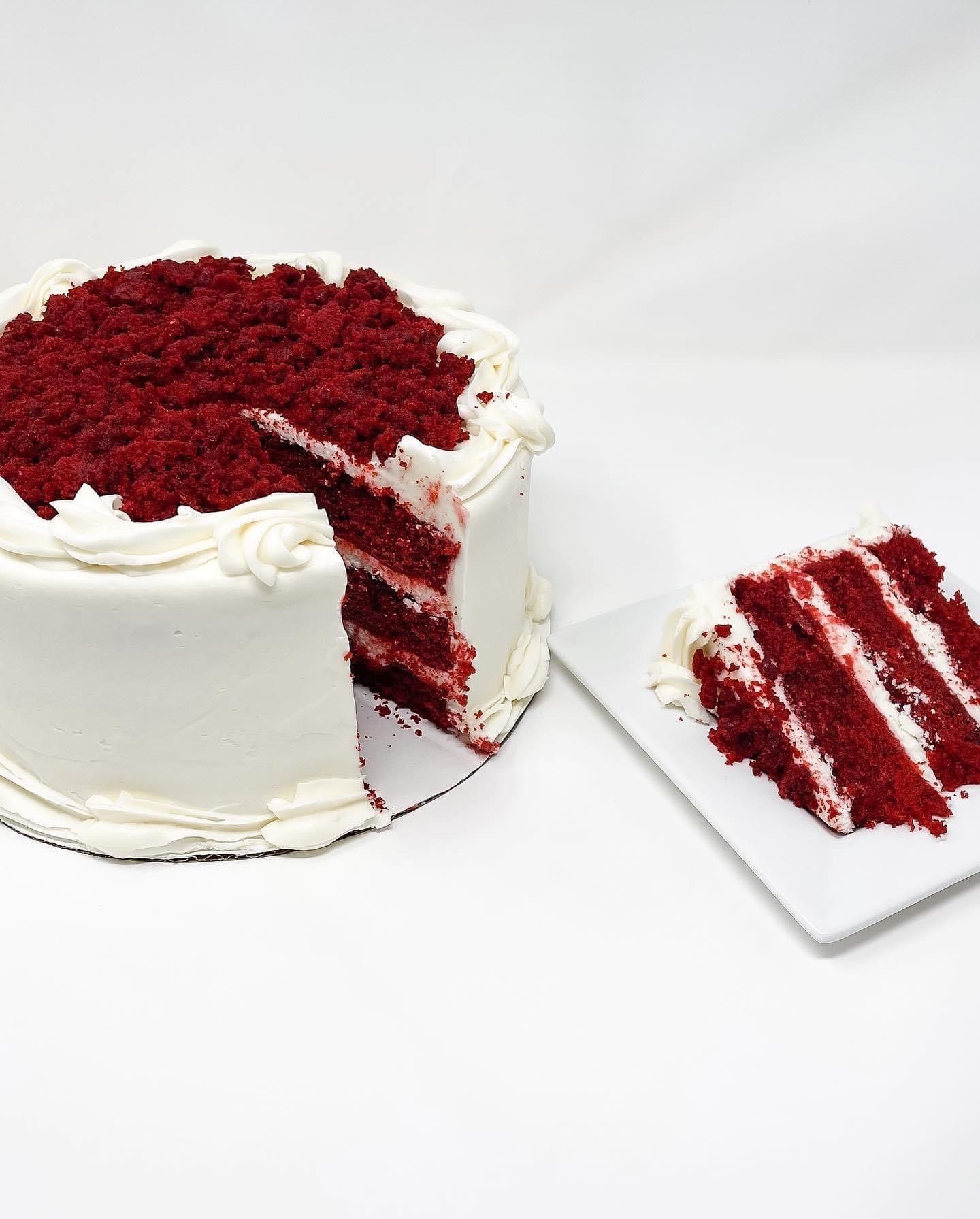 White frosted red velvet cake with a slice cut on a white plate, topped with red crumbs