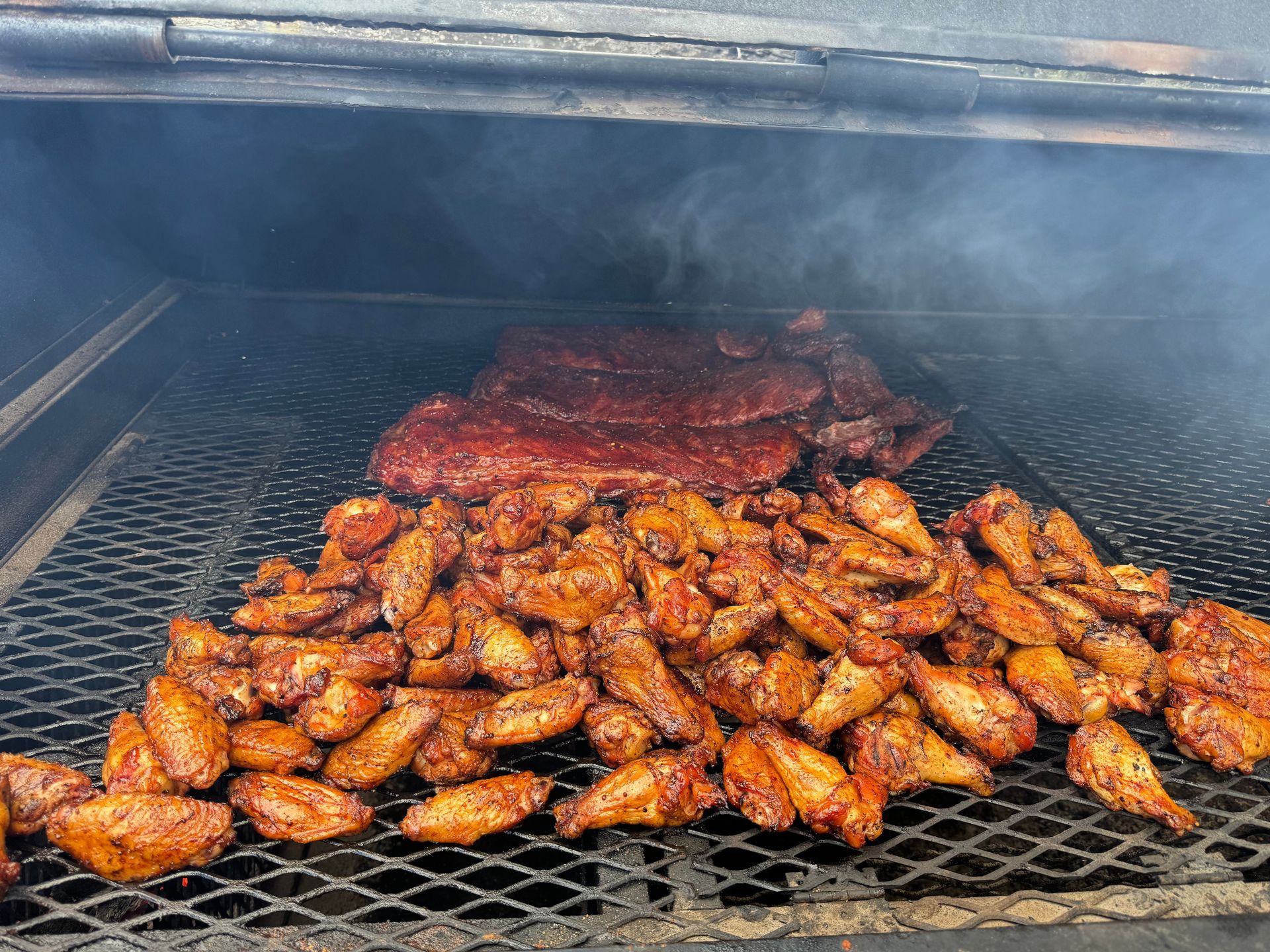 Chicken wings and ribs smoking on a grill inside a barbecue smoker, with smoke filling the chamber.