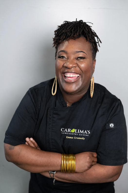 Smiling person in black shirt with gold jewelry, arms crossed against a light gray background