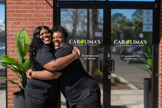 Sameka & 'Emma Two people hugging outside a brick storefront with glass doors and yellow lettering