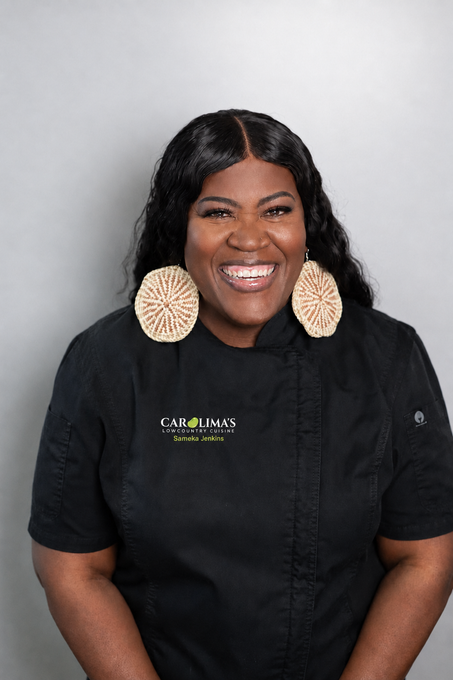 Smiling chef in black uniform with gold logo and large woven earrings, standing against a light background