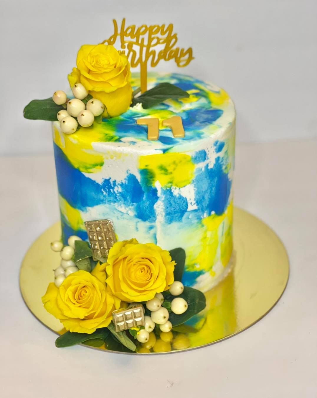 Yellow-and-blue birthday cake with roses and a “Happy Birthday” topper on a gold board