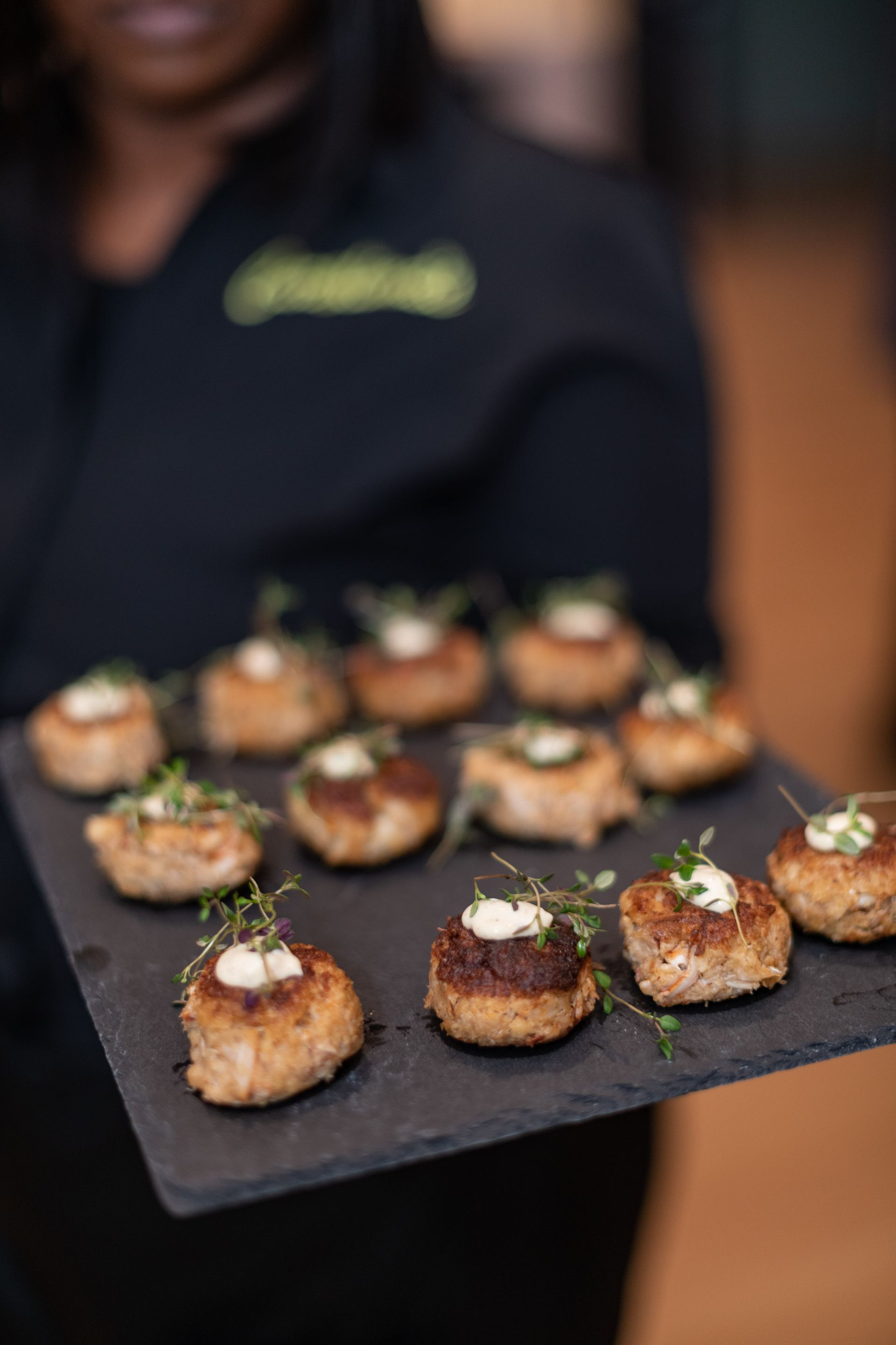 Tray of bite-sized appetizers on a black board, held by a person in a dark shirt.