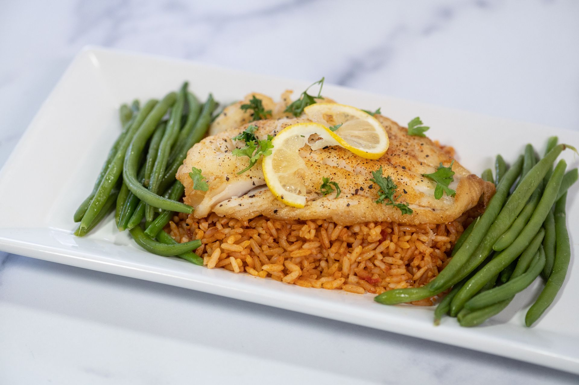 Plated lemon-garnished fish over rice with green beans on a white rectangular dish