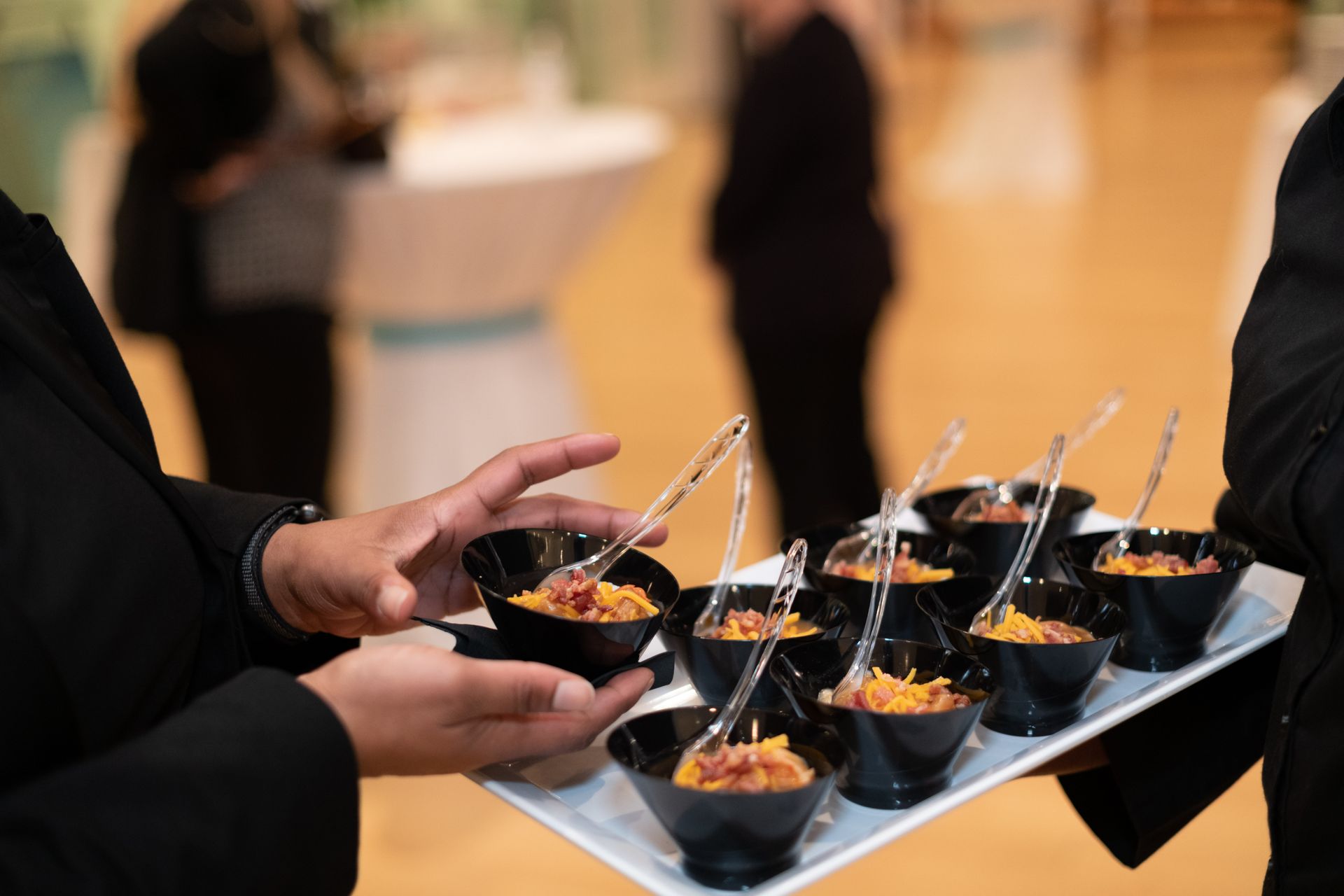Hands holding a tray of black appetizer cups with spoons at a catered event