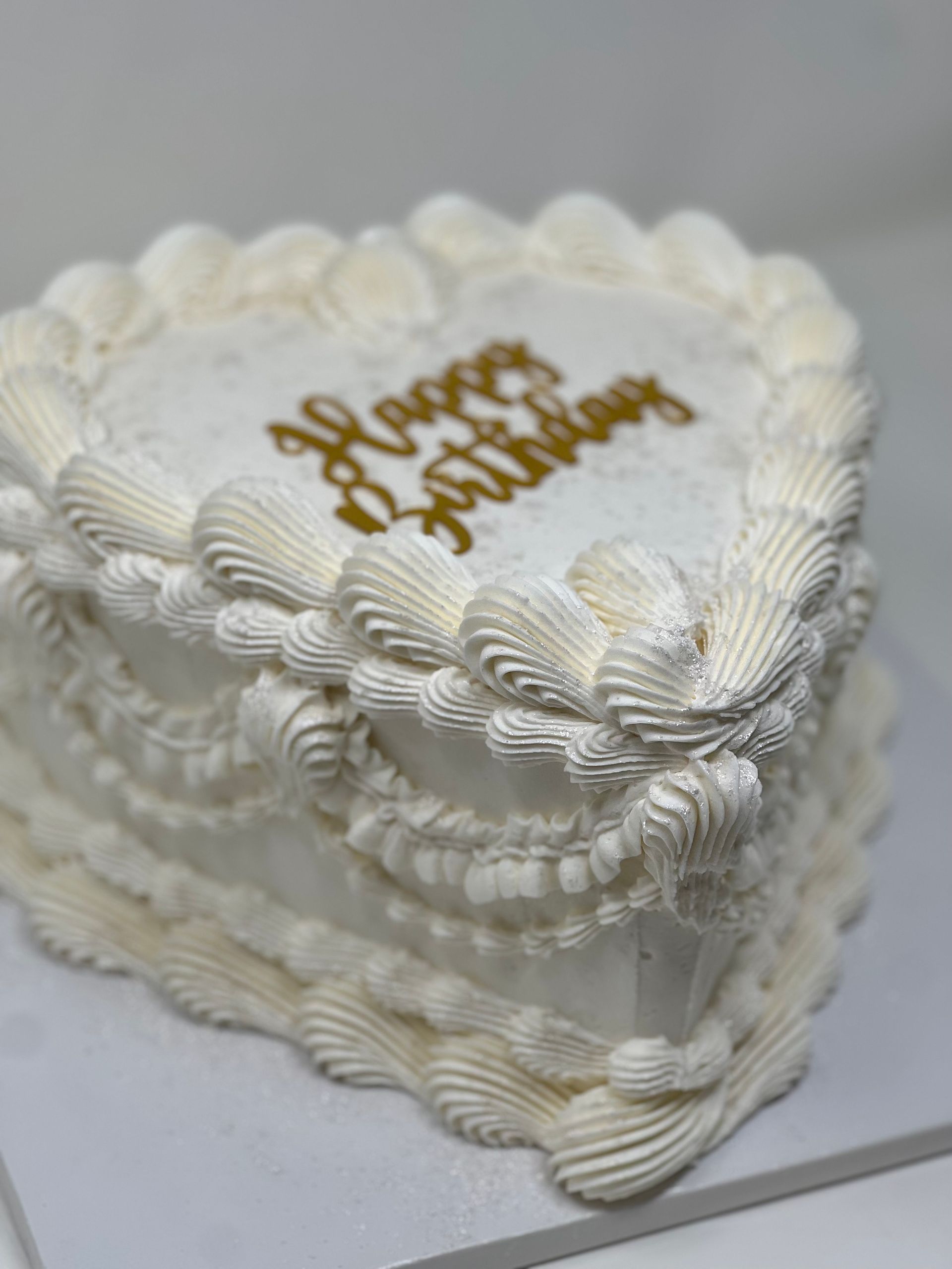 White heart-shaped birthday cake with piped frosting and gold “Happy Birthday” topper