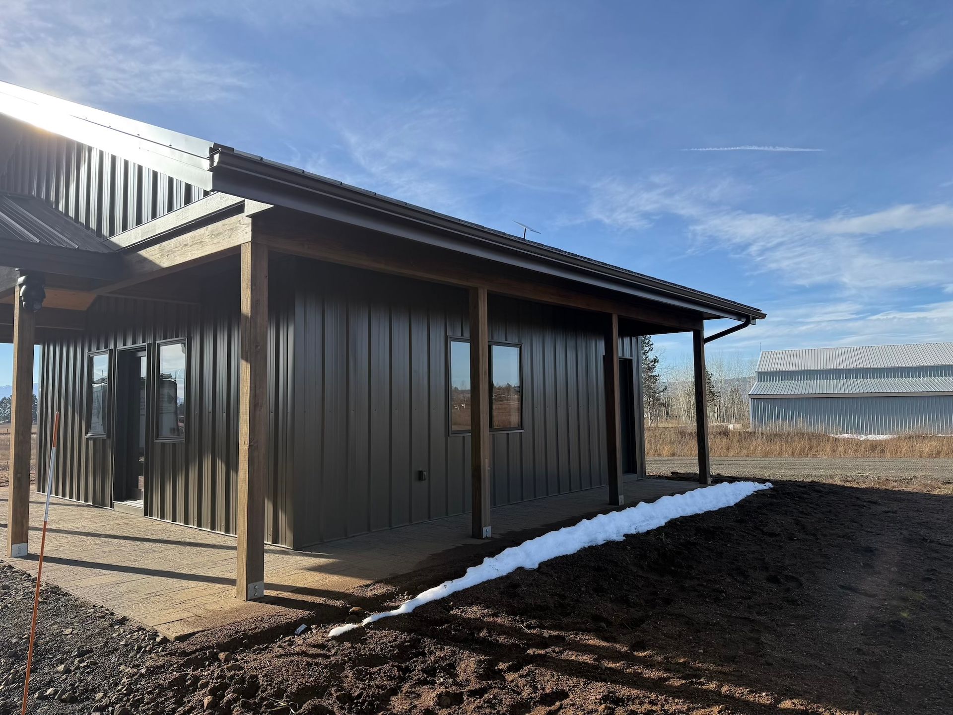 Small brown metal shed with corrugated roof on a gravel lot beside a greenhouse, with snow on the ground.